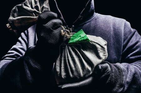Foto de Foto de aterrador hombre encapuchado sombreado sosteniendo una bolsa de banco de tela de monedas sobre fondo oscuro. - Imagen libre de derechos