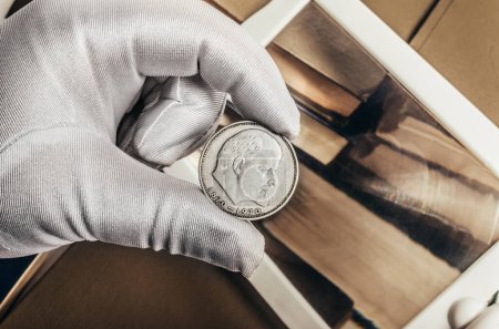 Photo for Photo of a person's male hand in white gloves holding a soviet 1970 ruble coin on stack of books and magnifying glass background. Numismatics hobby concept. - Royalty Free Image