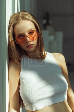 Téléchargez les photos : Jeune fille blonde avec des lunettes de soleil roses. Portrait intérieur avec lumière douce du soleil. Jolie babyface modèle - en image libre de droit