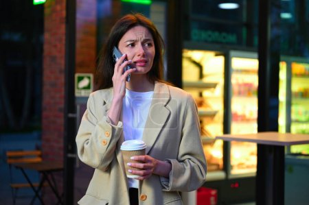 Photo for A beautiful woman dressed in casual attire is enjoying a cup of coffee in an urban setting. She appears to be deep in conversation as she speaks on her phone, with an expression that suggests she is both focused and content. - Royalty Free Image