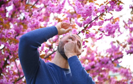 Foto de Un hombre está de pie cerca de un árbol con flores pero parece estar sufriendo de alergias. Se ve incómodo, frotándose la nariz y los ojos, y tal vez considerando tomar medicamentos para la alergia. - Imagen libre de derechos