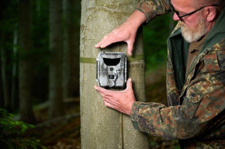 Foto de Hunter coloca una cámara de rastreo en un árbol en el bosque. Cámaras de sendero son a menudo utilizados por los cazadores para la fotografía automática o de vídeo de la vida silvestre en el bosque. - Imagen libre de derechos