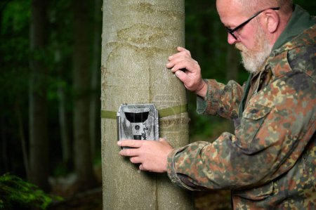 Foto de Hunter coloca una cámara de rastreo en un árbol en el bosque. Cámaras de sendero son a menudo utilizados por los cazadores para la fotografía automática o de vídeo de la vida silvestre en el bosque. - Imagen libre de derechos