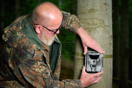 Téléchargez les photos : Hunter pose une caméra sur un arbre dans la forêt. Les caméras de piste sont souvent utilisées par les chasseurs pour la photographie automatique ou le tournage vidéo de la faune dans la forêt. - en image libre de droit