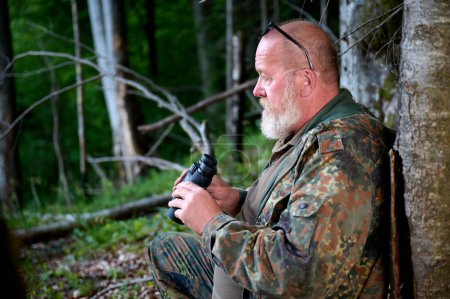 Foto de Cazador sénior de barba gris sosteniendo binoculares en las manos y un rifle sobre su hombro durante la caza, camina por el bosque, mira a través de binoculares y observa la naturaleza. Concepto de caza y viajes - Imagen libre de derechos