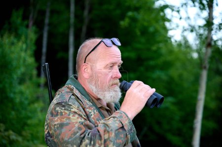 Foto de Hombre cazador de viajeros con prismáticos en sus manos y un rifle sobre su hombro camina por el bosque, mira a través de prismáticos y observa la naturaleza - Imagen libre de derechos