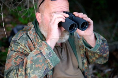 Téléchargez les photos : Homme chasseur de voyageurs avec des jumelles dans ses mains et un fusil sur son épaule marche à travers la forêt, regarde à travers les jumelles et observe la nature - en image libre de droit
