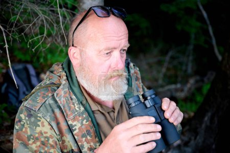 Téléchargez les photos : Homme chasseur de voyageurs avec des jumelles dans ses mains et un fusil sur son épaule marche à travers la forêt, regarde à travers les jumelles et observe la nature - en image libre de droit