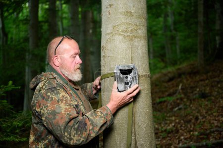 Photo for Hunter sets a trail camera on a tree in the forest. Trail cameras are often used by hunters for automatic photography or video shooting of wildlife in the forest. - Royalty Free Image