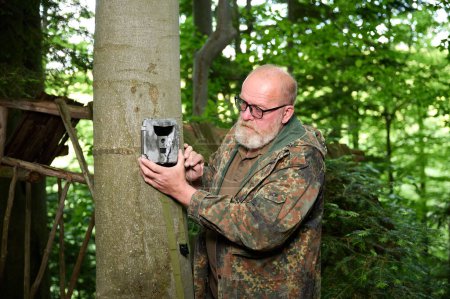 Téléchargez les photos : Hunter pose une caméra sur un arbre dans la forêt. Les caméras de piste sont souvent utilisées par les chasseurs pour la photographie automatique ou le tournage vidéo de la faune dans la forêt. - en image libre de droit