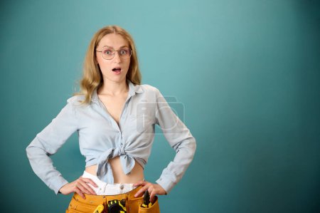 Photo for Portrait of surprised young female worker with tool belt isolated on background. Pretty caucasian female with tools planning new project. Portrait in studio. Copy space. - Royalty Free Image
