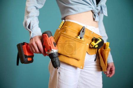 Photo for Young female worker with tool belt holding an electric screwdriver in hands isolated on background. Pretty caucasian female with tools planning new project. Portrait in studio. - Royalty Free Image