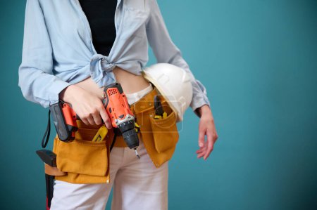 Photo for Young female worker with tool belt holding an electric screwdriver in hands isolated on background. Pretty caucasian female with tools planning new project. Portrait in studio. - Royalty Free Image