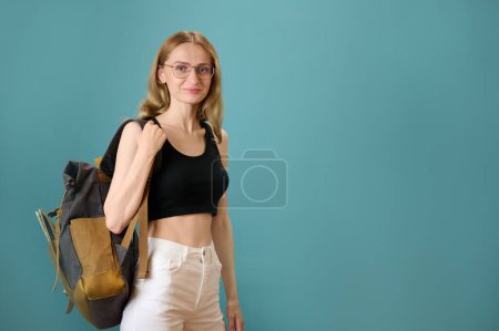 Foto de Joven hipster bastante femenino con mochila vintage aislado en el fondo. Atractiva estudiante femenina en gafas, top corto negro y pantalones blancos. Mujer sonriente mostrando expresiones. Viaje con mochila - Imagen libre de derechos