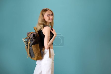 Photo for Young pretty female hipster with vintage backpack isolated on background. Attractive female student in glasses, short black top and white pants. Smiling woman showing expressions. Travel with backpack - Royalty Free Image