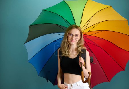 Photo for Happy caucasian female in black top and white pants under rainbow colored umbrella posing at studio background, free copy space - Royalty Free Image