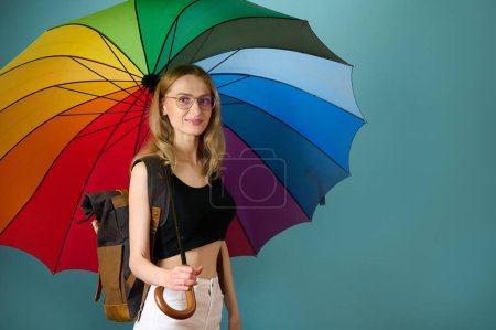 Téléchargez les photos : Joyeuse femme caucasienne en haut noir et pantalon blanc sous parapluie coloré arc-en-ciel posant en arrière-plan studio, espace de copie gratuit - en image libre de droit