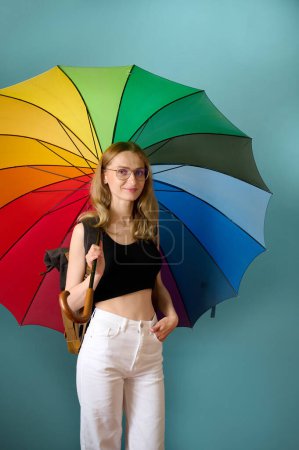 Téléchargez les photos : Joyeuse femme caucasienne en haut noir et pantalon blanc sous parapluie coloré arc-en-ciel posant en arrière-plan studio, espace de copie gratuit - en image libre de droit