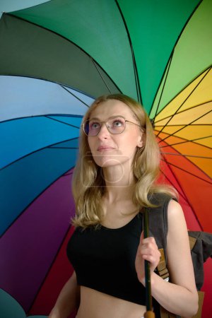 Photo for Happy caucasian female in black top and white pants under rainbow colored umbrella posing at studio background, free copy space - Royalty Free Image