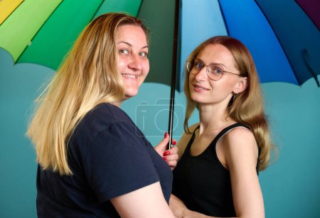 Foto de Dos jóvenes lesbianas felices se abrazan bajo un colorido paraguas. Atractivo apoyo femenino, pareja de lesbianas de pie y abrazos. - Imagen libre de derechos