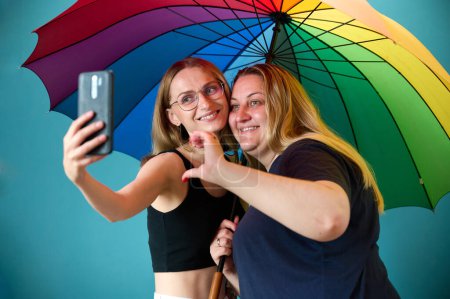 Foto de Dos jóvenes lesbianas felices se abrazan bajo un colorido paraguas. Atractivo apoyo femenino, abrazo de pareja lesbiana. Dos amigas tomando selfies bajo paraguas coloreadas en colores de bandera LGBTQ. - Imagen libre de derechos