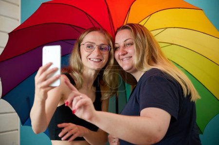 Photo for Two happy young lesbian girls hug under a colorful umbrella. Attractive carring female support, lesbian couple hugging. Two female friends taking selfies under umbrella colored in LGBTQ flag colors. - Royalty Free Image