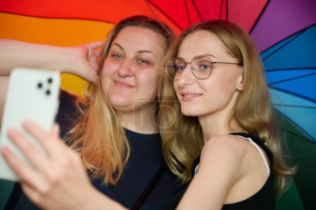 Téléchargez les photos : Deux jeunes filles lesbiennes heureuses s'embrassent sous un parapluie coloré. Attrayant soutien féminin attentionné, couple lesbien câlin. Deux amies prenant des selfies sous un parapluie coloré aux couleurs du drapeau LGBTQ. - en image libre de droit