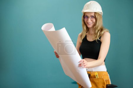 Photo for Young architect girl with protective helmet holding a house plan in hands isolated on background. Pretty caucasian female architect are planning new project. People work on architecture project - Royalty Free Image