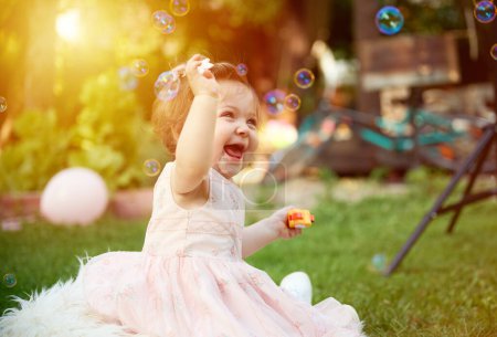 Photo for Adorable girl on the grass in the garden. Close up portrait. Happy little girl in summer scenery. Sweet small kid outdoors. A child plays with soap bubbles. - Royalty Free Image