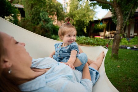 Téléchargez les photos : Maman et sa pâte jouant dans le jardin, allongés dans un hamac. Belle famille posant à l'extérieur. Bonne petite fille dans les paysages d'été. Doux petit enfant à l'extérieur. - en image libre de droit