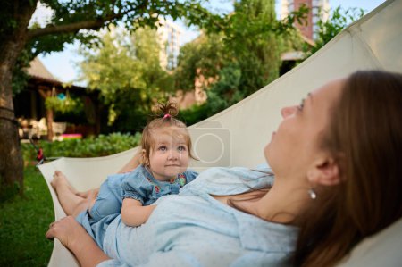 Photo for Mommy and her doughter playing in the garden, lying in hammock. Lovely family posing outdoors. Happy little girl in summer scenery. Sweet small kid outdoors. - Royalty Free Image