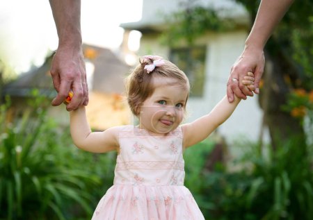 Photo for Lovely family walking holding hands, adopted child being supported by loving parents. Girl holds hands of her parents and jumps. Cropportrait of the cute adorable small girl in dress. - Royalty Free Image