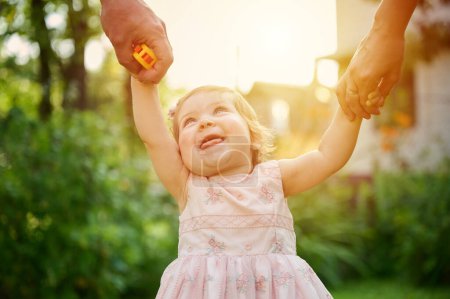 Photo for Lovely family walking holding hands, adopted child being supported by loving parents. Girl holds hands of her parents and jumps. Cropportrait of the cute adorable small girl in dress. - Royalty Free Image