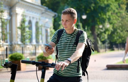 Téléchargez les photos : Jeune homme sportif dans un T-shirt rayé enregistre un tour sur un scooter électrique. Louer un scooter électrique dans la ville. Transport urbain électrique. Numérisation de la main de l'homme avec un téléphone mobile le code QR - en image libre de droit