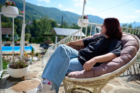 Téléchargez les photos : Jolie fille se détendre sur la station balnéaire dans les montagnes des Carpates. Vue latérale d'une jeune femme assise sur une chaise et regardant un beau paysage. Fille avec en vêtements décontractés sur fond de montagnes. - en image libre de droit