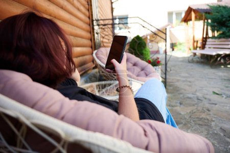 Téléchargez les photos : Jolie fille se détendre sur la station balnéaire dans les montagnes des Carpates. Vue arrière de la jeune femme assise sur une chaise et regardant le téléphone. Fille avec en vêtements décontractés sur fond de montagnes. - en image libre de droit