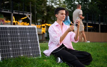 Téléchargez les photos : Jeune femme recharge son téléphone par panneau solaire. Jolie femelle en vêtements décontractés assis sur l'herbe et branchant chargeur dans son téléphone. Concept d'énergie renouvelable. - en image libre de droit