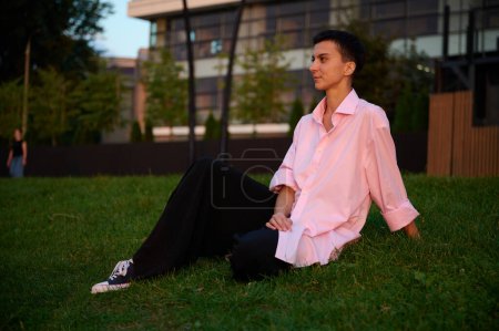 Téléchargez les photos : Jeune fille aux cheveux courts est assis sur une pelouse verte profiter du coucher du soleil.Jolie femelle en vêtements décontractés profiter de la vie en plein air. Zone verte dans l'espace urbain. - en image libre de droit