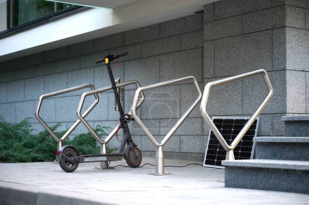 Téléchargez les photos : Scooter électrique garé dans un parking moderne près de la maison avec recharge à partir d'un panneau solaire - en image libre de droit