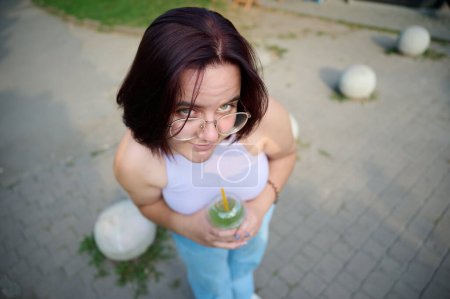 Photo for Pretty girl drinking lemonade at sunny day. Young female squinting her eyes because of sunlight. Positive girl drinking and relaxing outside. Close up top view - Royalty Free Image