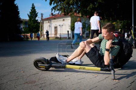 Photo for Careless man felt down from electric scooter. Young male lying on back after falling from his vehicle. Mused male collided with a car and hurted his knees. - Royalty Free Image