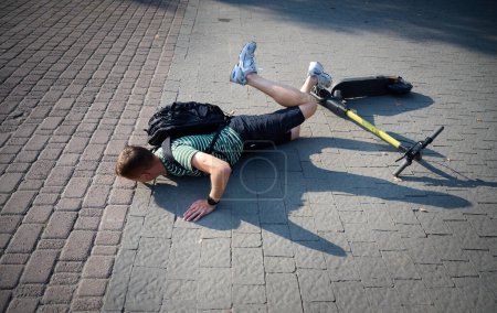 Photo for Careless man felt down from electric scooter. Young male lying on back after falling from his vehicle. Mused male collided with a car and hurted his back. - Royalty Free Image
