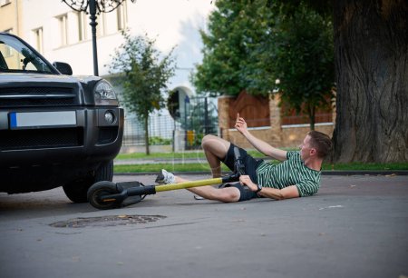Photo for Careless man felt down from electric scooter. Young male lying on back after falling from his vehicle. Mused male collided with a car and hurted his back. - Royalty Free Image