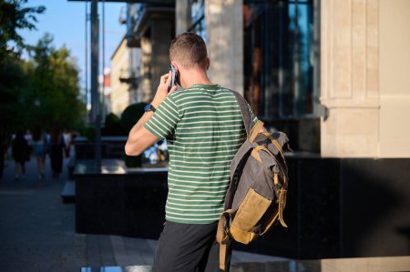 Téléchargez les photos : Jeune homme sportif en T-shirt rayé marchant dans la rue avec un smartphone à la main et un sac à dos branché pendu sur son épaule, profitant d'une sortie urbaine tranquille. - en image libre de droit