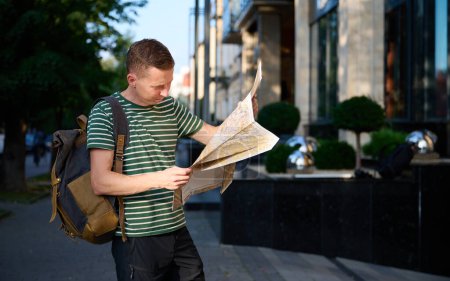 Photo for Young traveller in a striped T-shirt walking on the street with a map and smartphone in hand and a trendy hipster's backpack slung over his shoulder, enjoying a leisurely urban outing. - Royalty Free Image