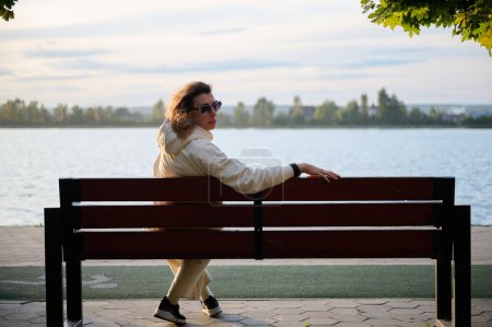 Photo for View from the back woman in a modern sun glasses sitting on the wooden bench in a park near a lake enjoying the sunset.Stylish girl in light sports clothes sitting and relaxing on the beach at sunset - Royalty Free Image