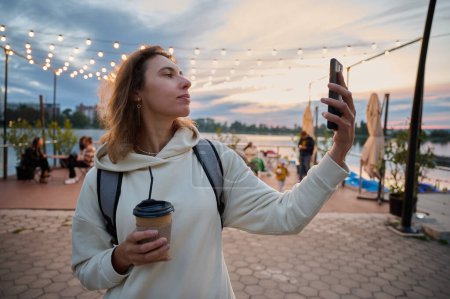 Foto de Retrato de una mujer bonita caminando por la playa, bebiendo té, tomando una selfie y navegando por las redes sociales por teléfono. Hermosa puesta de sol en la playa y hermosa iluminación artificial en un café de playa - Imagen libre de derechos