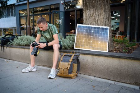Photo for Traveling photographer using solar panel to charge his camera. Young traveler waiting for his camera to charge. Positive male relaxing after long walk. - Royalty Free Image