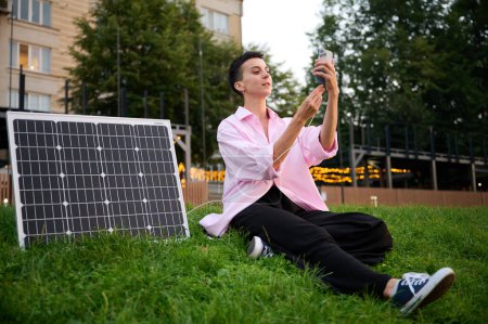 Foto de Mujer joven cargando su teléfono por el panel solar. Mujer bonita con ropa casual sentada en la hierba y cargador de enchufe en su teléfono. Concepto de energía renovable. - Imagen libre de derechos