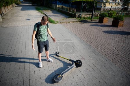 Photo for Careless man felt down from electric scooter. Young male standing next to a broken electric scooter after an accident after falling from his vehicle. Mused male collided with a car and hurted his back - Royalty Free Image